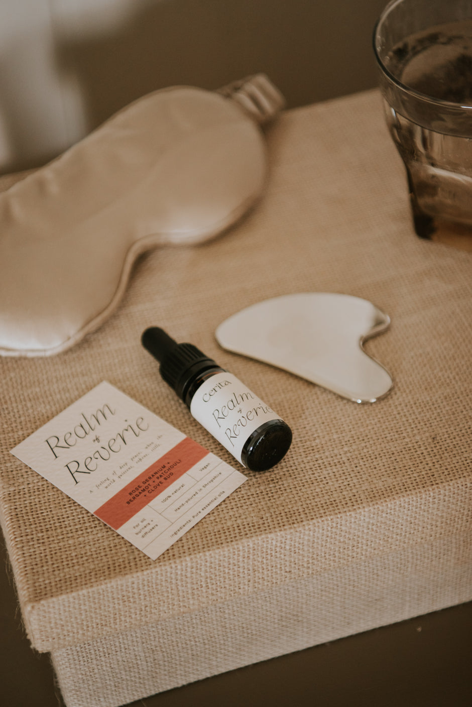 bottle of realm of reverie essential oil blend by Cerita on bedside table with silk sleep mask, gua sha and glass of water