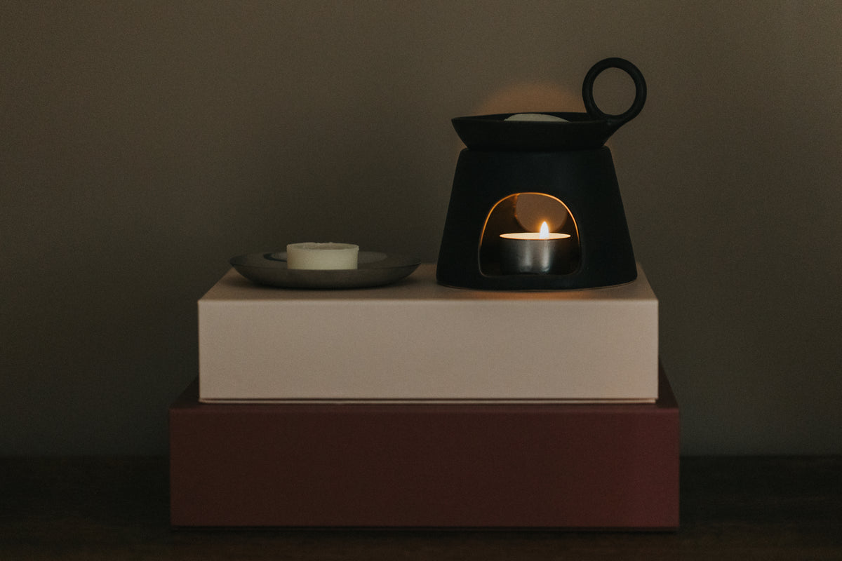 wax melt on silver dish next to black oil burner with lit tealight, both on top of stacked gift boxes