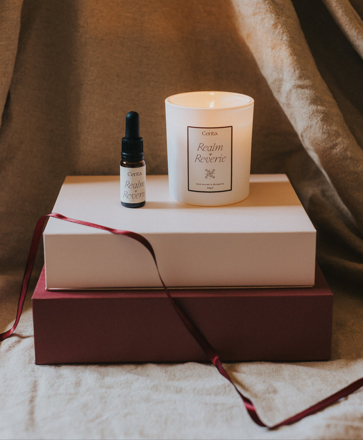 bottle of essential oil blend and lit aromatherapy candle on stacked gift boxes with draped ribbon