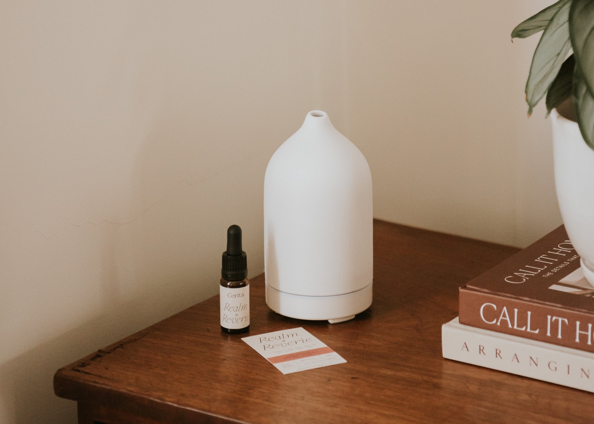 bottle of realm of reverie essential oil blend by Cerita next to white ceramic diffuser, books and plant on wooden surface